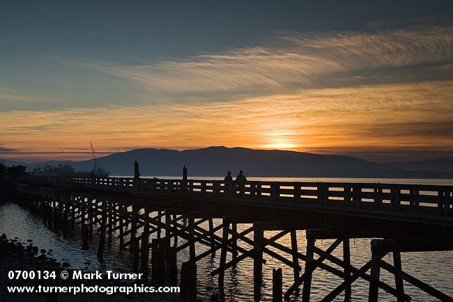 0700134 Sunset over South Bay Trail trestle over edge of Bellingham Bay w/ silhouettes of people walking, Lummi Island bkgnd. Bellingham, Boulevard Park, WA. © Mark Turner